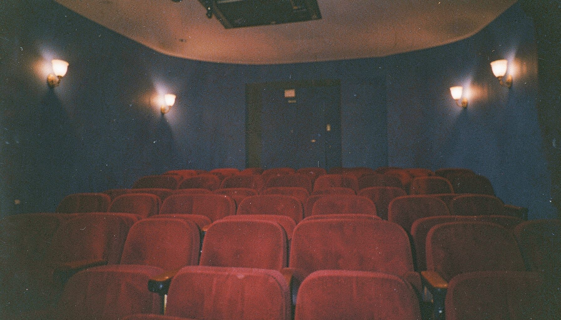 A grainy analog film photograph of a warmly lit cinema auditorium with deep red velvet seats and navy blue walls, soft amber glow from wall sconces, moody and atmospheric