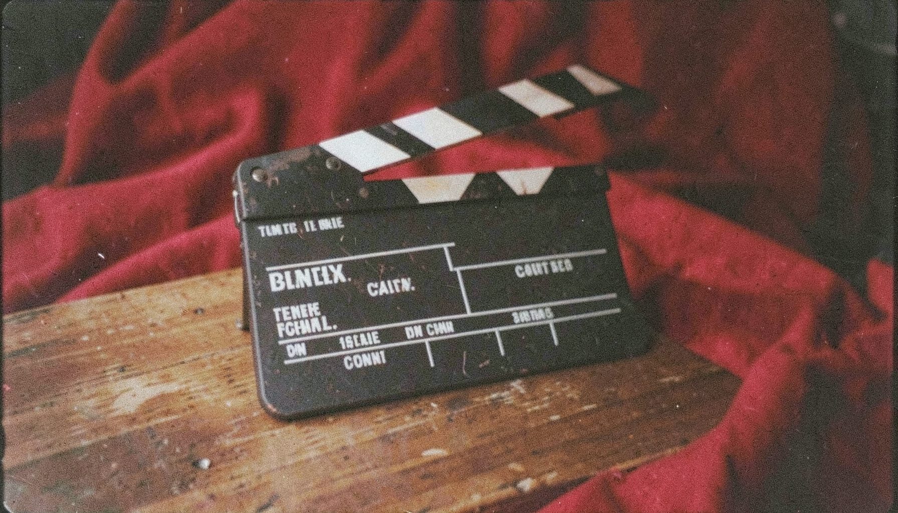 A grainy analog film photograph of a clapperboard on a wooden table against deep red fabric, cinematic atmosphere with soft shadows