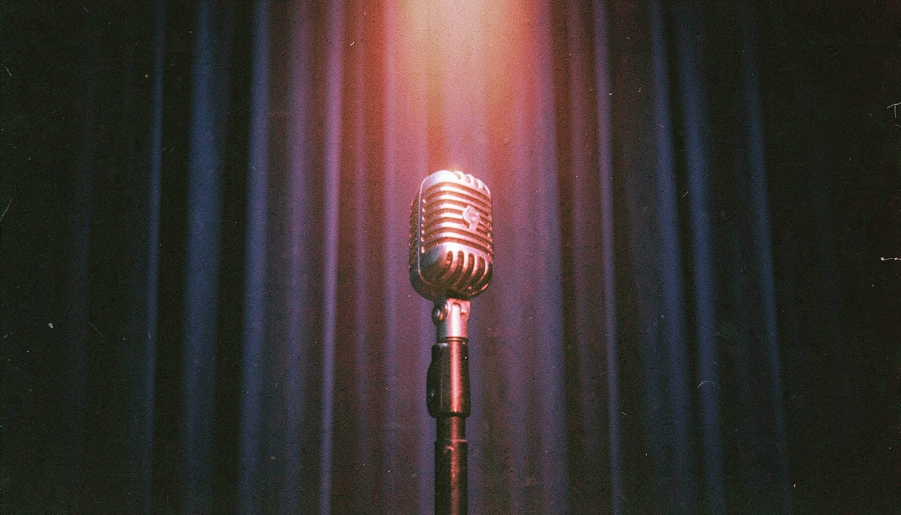 A grainy analog film photograph of a vintage microphone on a dark stage with navy blue curtains and warm spotlight, intimate performance mood