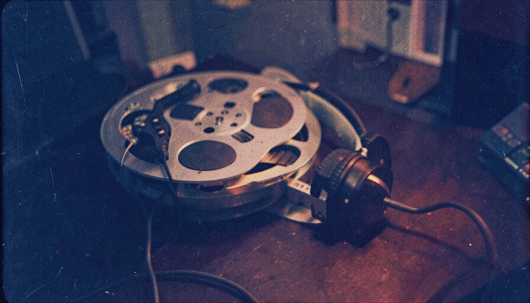 A grainy analog film photograph of a film reel and headphones on a dark desk, navy blue tones with warm amber accent light