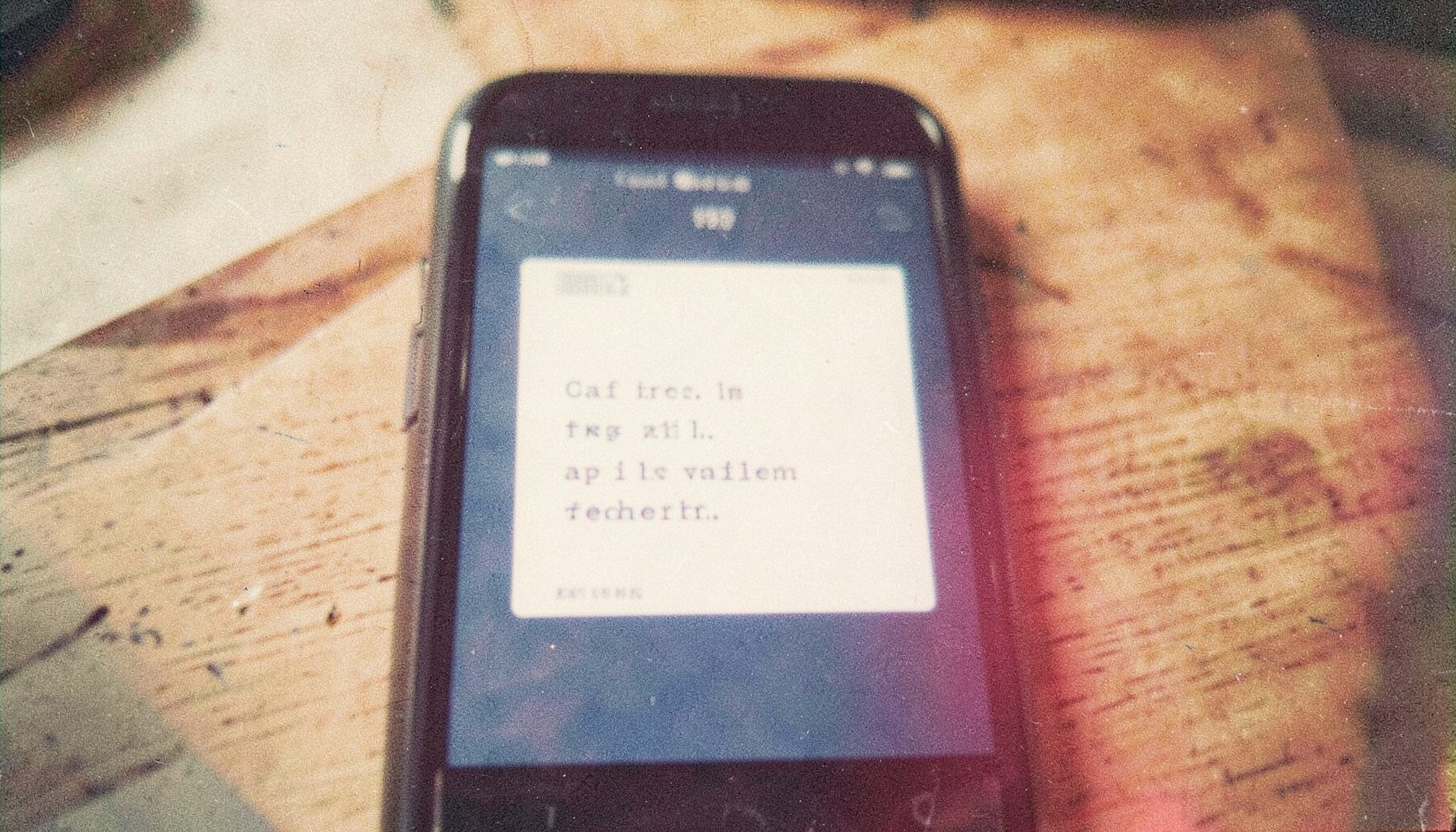 A grainy analog film photograph of a smartphone screen displaying text on a wooden surface, soft natural light, muted navy and cream tones