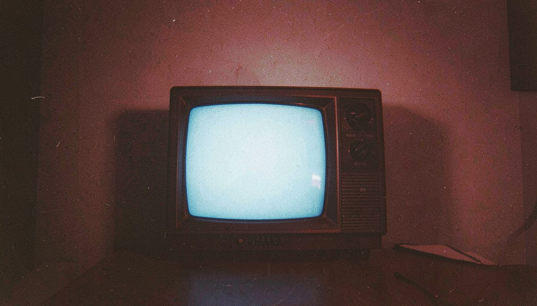 Grainy film photograph of a vintage television set glowing with cool blue light in a dark room, casting soft shadows on the wall behind it.