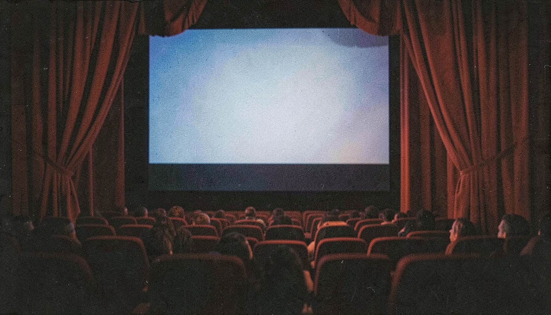 Analog film photograph with grain showing a cinema screen in a dim theater, deep red curtains framing the edges and soft blue light spilling across empty seats.