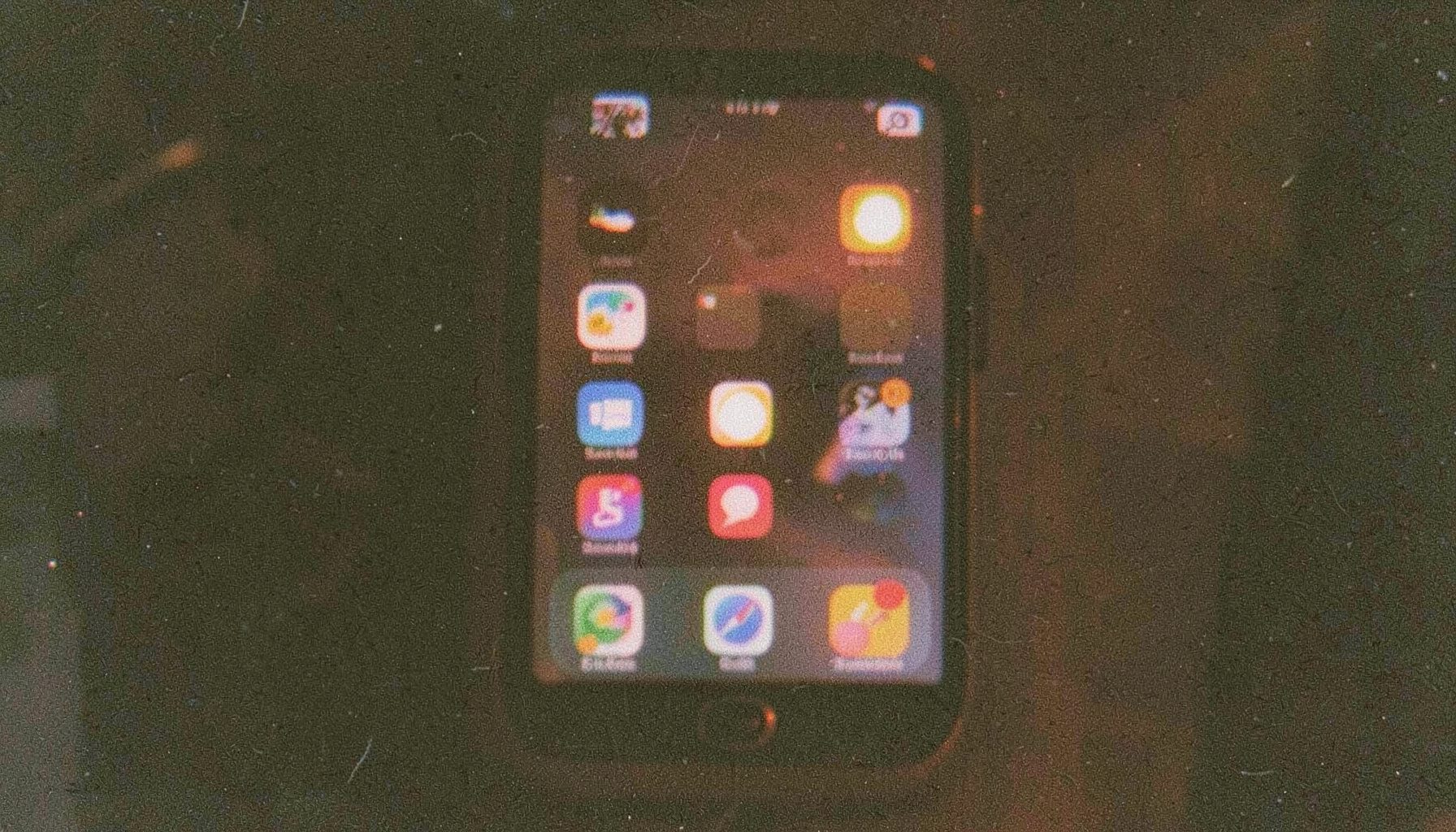 Grainy analog film photo of a smartphone screen displaying colorful social media icons against a dark background with warm amber reflections.