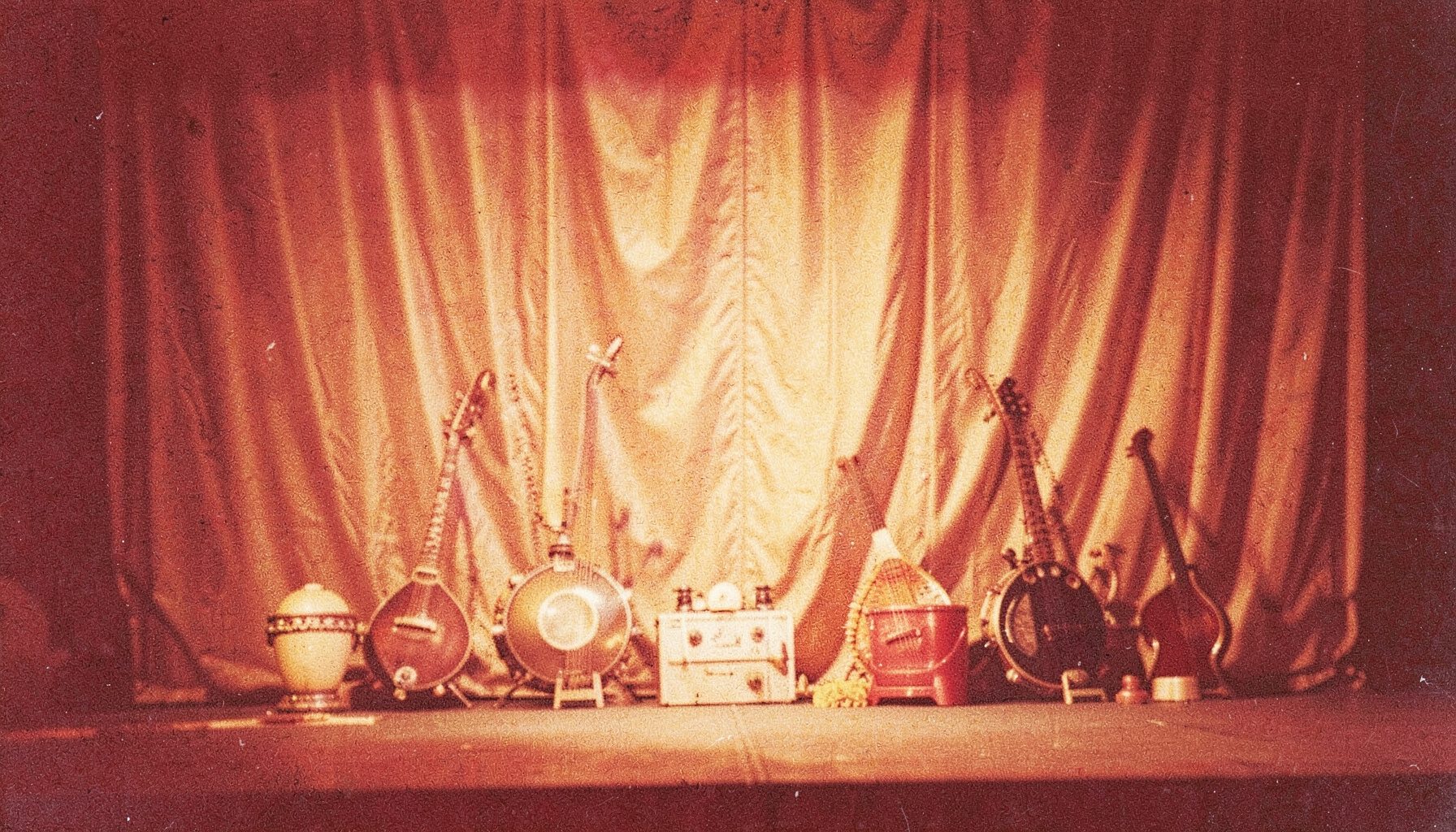 Warm-toned analog film photograph with visible grain showing a softly lit stage with traditional Indian instruments resting against a draped backdrop in deep amber and gold hues.