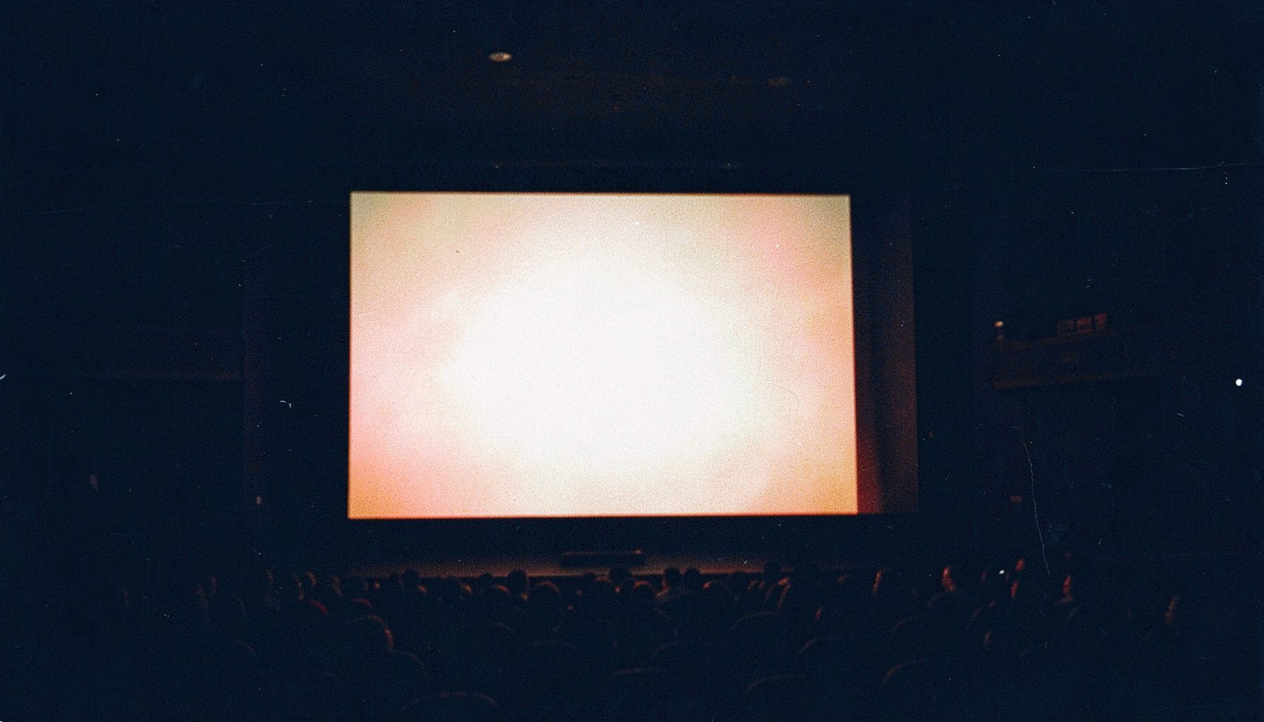 A grainy analog film photograph of a cinema screen glowing in a darkened theater, deep navy shadows and warm amber highlights, moody and atmospheric