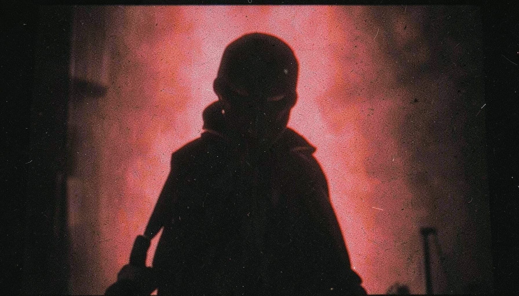 Grainy analog film still in muted reds and blacks, evoking an action-comic aesthetic with a masked figure silhouette.