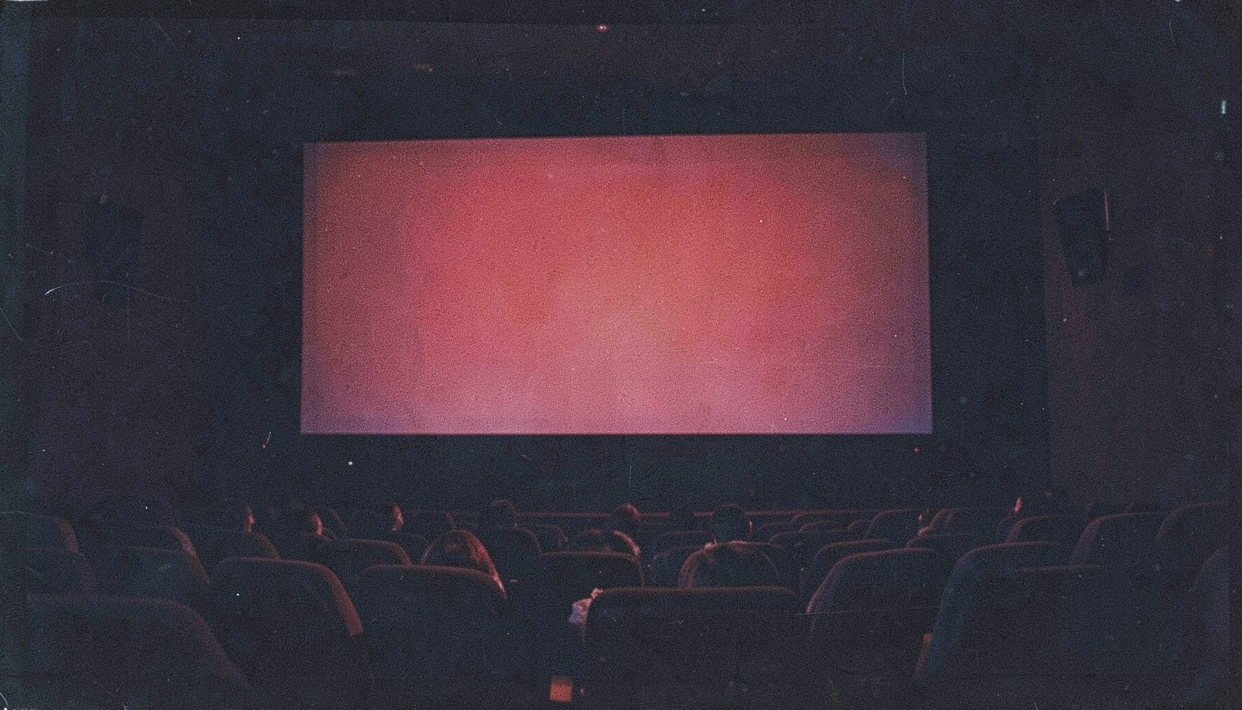 A dark, moody film-still style photograph with visible grain, deep red and navy tones, showing a cinema screen glow against shadowed theater seats.